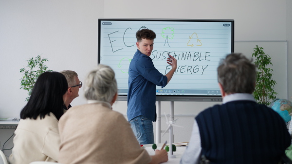 A young teacher writing on whiteboard during alternative energy lecture with group of senior students in classroom.