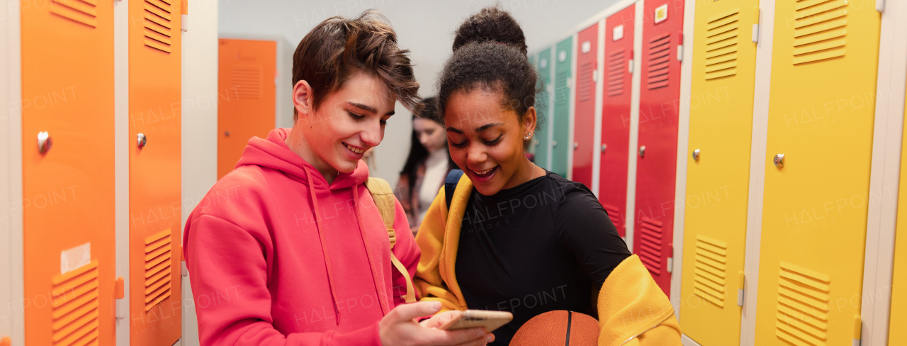 Young high school students standing near locker in campus hallway talking and using a smartphone.