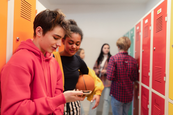 Young high school students standing near locker in campus hallway talking and using a smartphone.