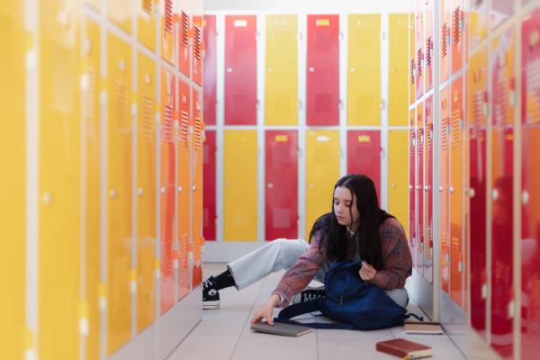 A teenage student sitting in corridor near colorful lockers adn packing book to backpack in campus hallway, back to school concept.