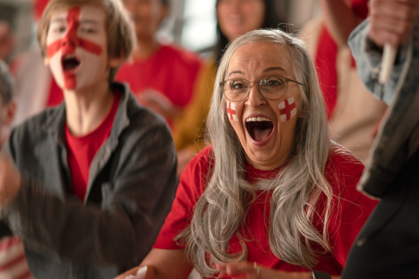 An excited senior woman football fan supproting English national team in live soccer match at stadium.