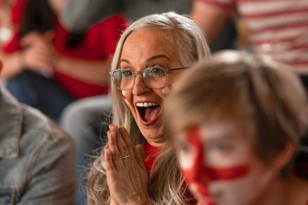 An excited senior woman football fan supproting English national team in live soccer match at stadium.