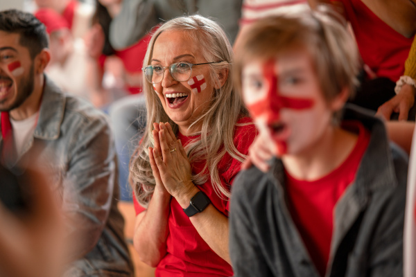 An excited senior woman football fan supproting English national team in live soccer match at stadium.