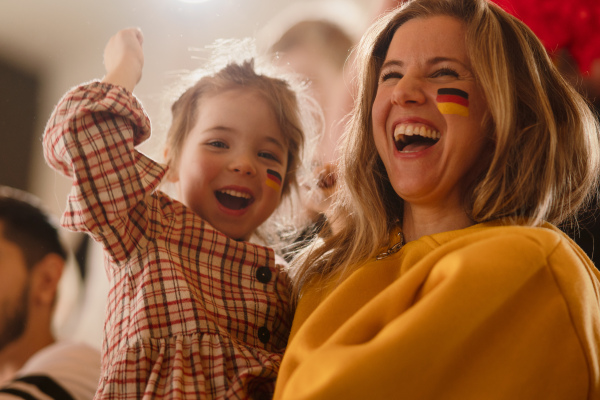 Excited football fans, mother with little daughter, supproting a German national team in live soccer match at stadium.