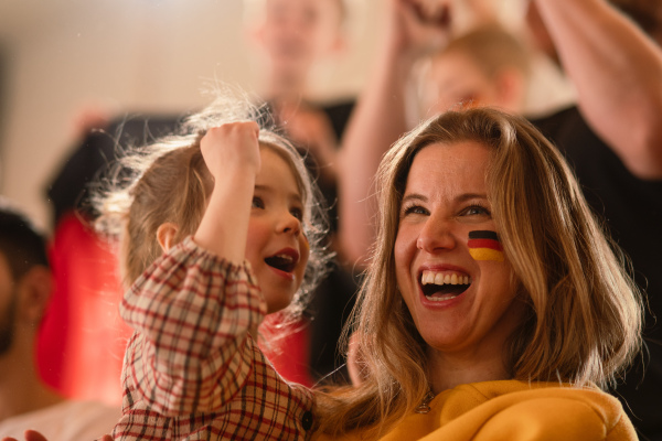 Excited football fans, mother with little daughter, supproting a German national team in live soccer match at stadium.