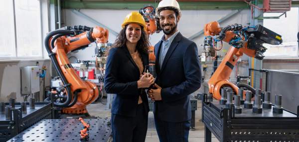 A portrait of man engineer with his female collegue standing in factory, in front of robotic arms.