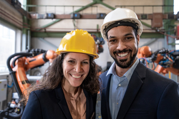 A chief engineer and project manager in modern industrial factory with robot arms looking at camera.