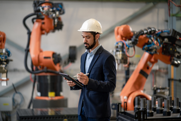 An automation engineer uses tablet for programming robotic arm in factory.