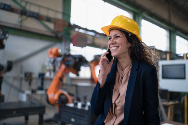 An automation female engineer with tablet for programming robotic arm calling on cellphone and walking through factory.