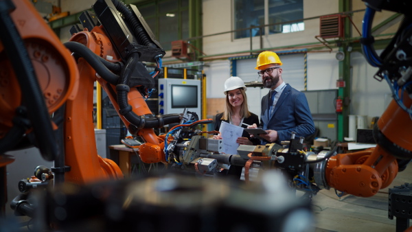 A chief engineer and project manager in modern industrial factory with robot arms talking and planning optimization of production.