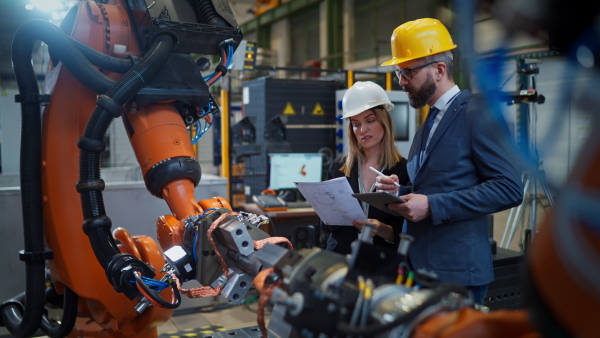 A chief engineer and project manager in modern industrial factory with robot arms talking and planning optimization of production.