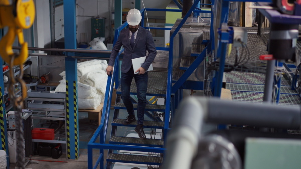 A chief Engineer in the hard hat walks through industrial factory while holding tablet.