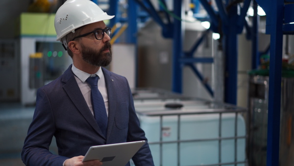 A chief Engineer in the hard hat walks through industrial factory while holding tablet.