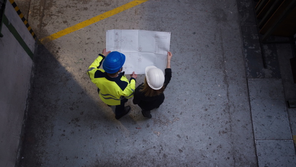 A high angle view of anager supervisor engineer and industrial worker in uniform discussing blueprints in large metal factory hall.