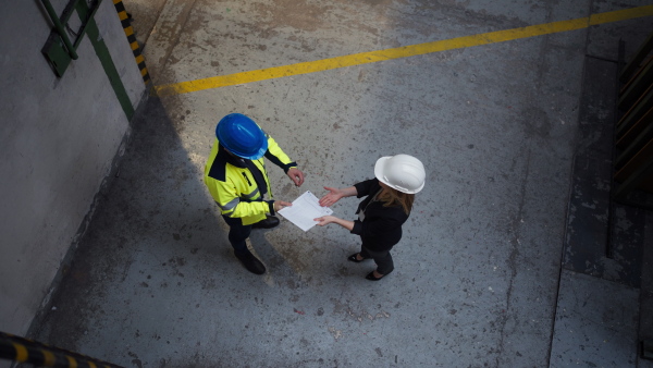 A high angle view of anager supervisor engineer and industrial worker in uniform discussing blueprints in large metal factory hall.