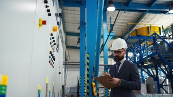 A chief Engineer in the hard hat walks through industrial factory while holding tablet.