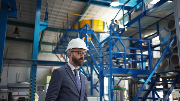 A chief Engineer in the hard hat walks through industrial factory while holding tablet.