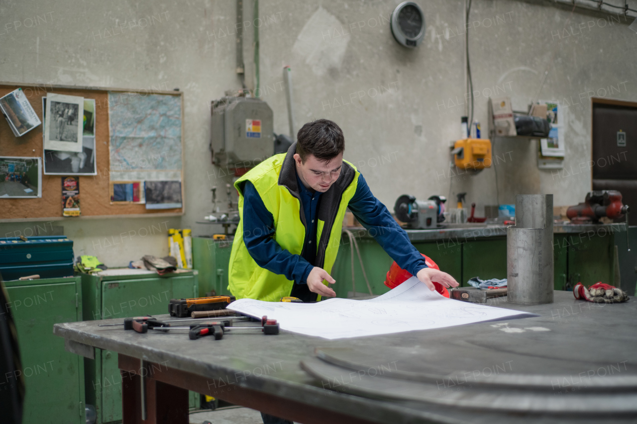 A young man with Down syndrome looking at blueprints when working in industrial factory, social integration concept.