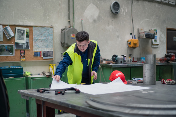 A young man with Down syndrome looking at blueprints when working in industrial factory, social integration concept.