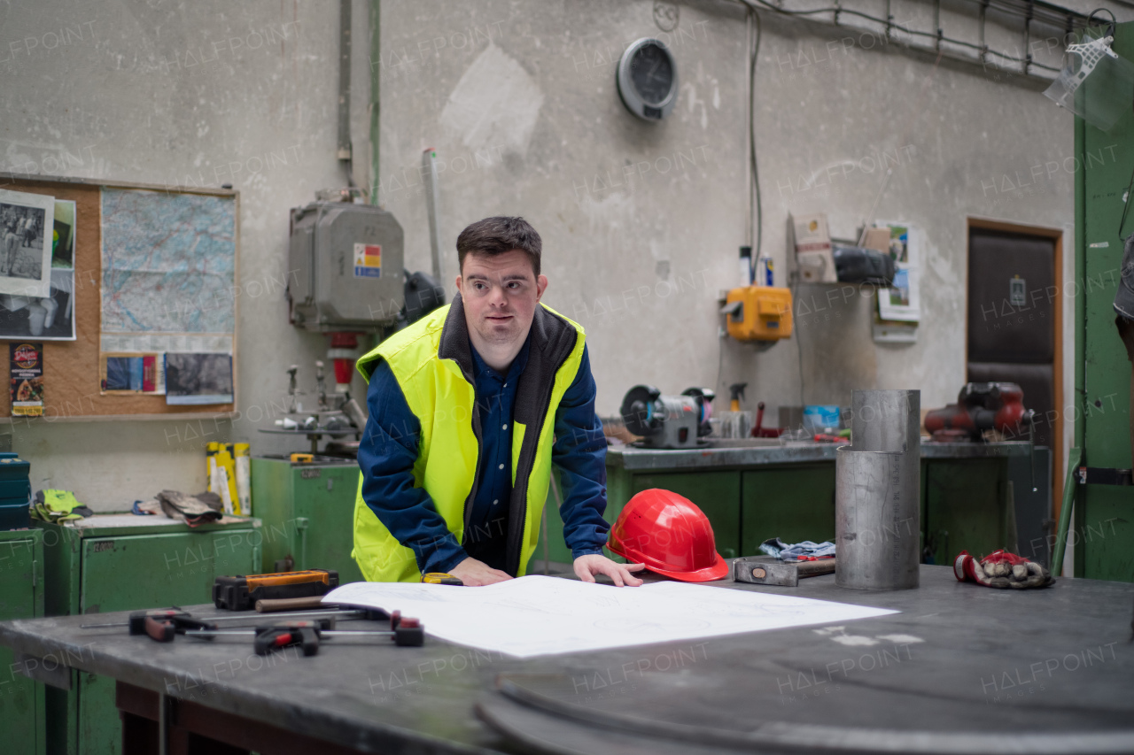 A young man with Down syndrome looking at blueprints when working in industrial factory, social integration concept.