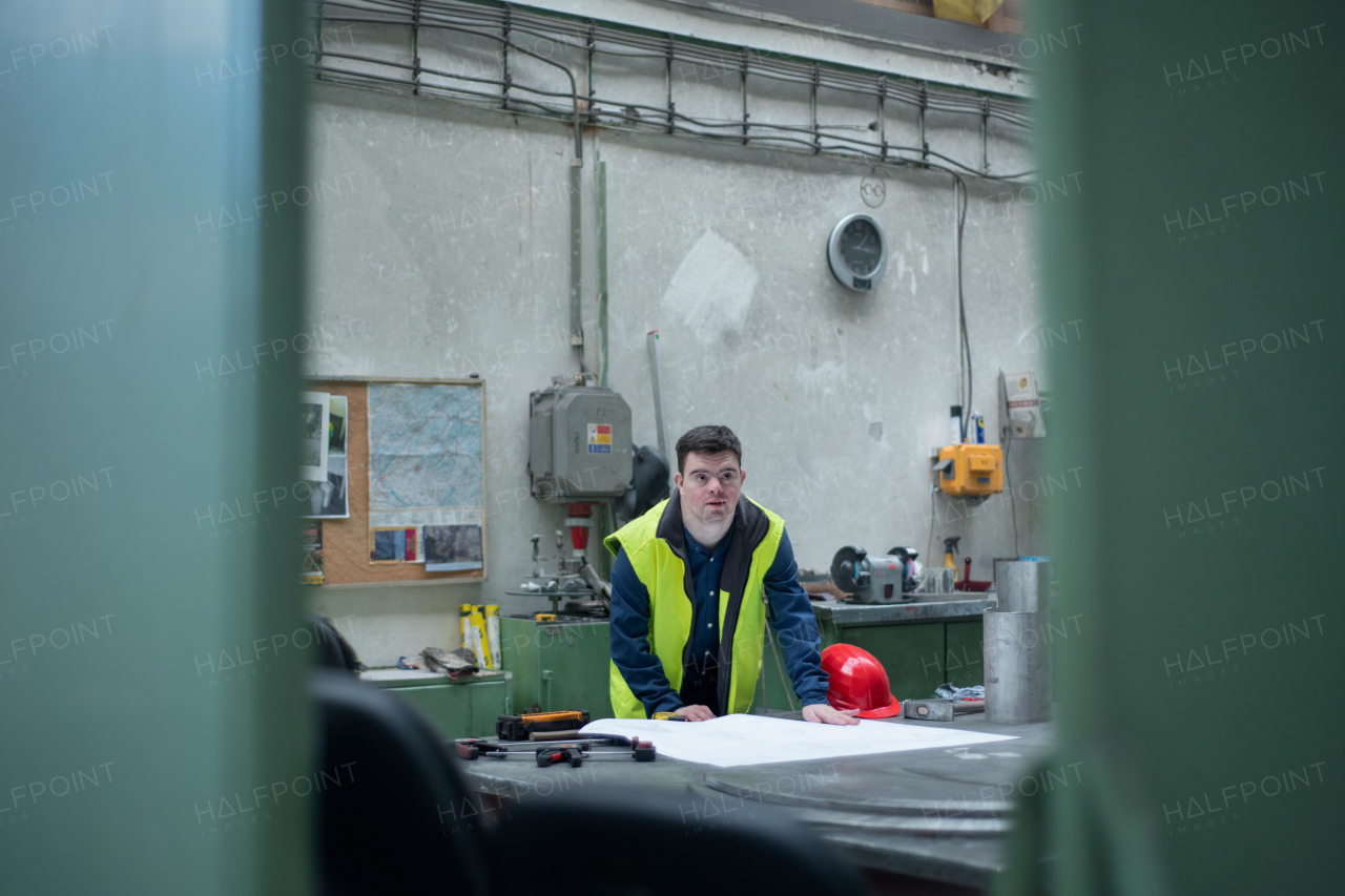 A young man with Down syndrome looking at blueprints when working in industrial factory, social integration concept.