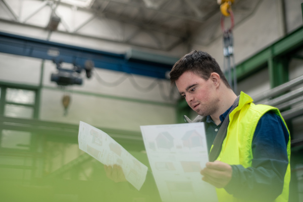 A young man with Down syndrome looking at blueprints when working in industrial factory, social integration concept.