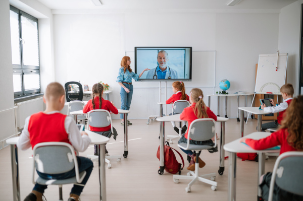 A rear view,class of pupils sitting, listening to teacher and wathing online lecture with doctor in classroom