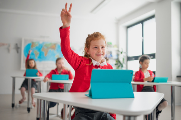 A happy schoolgirl with hands up using digital tablet during lesson in classroom at primary school.