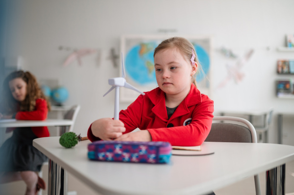 A Down syndrome schoolgirl with model of wind turbine learning about eco-friendly renewable sources of energy in class at school, integration concept.