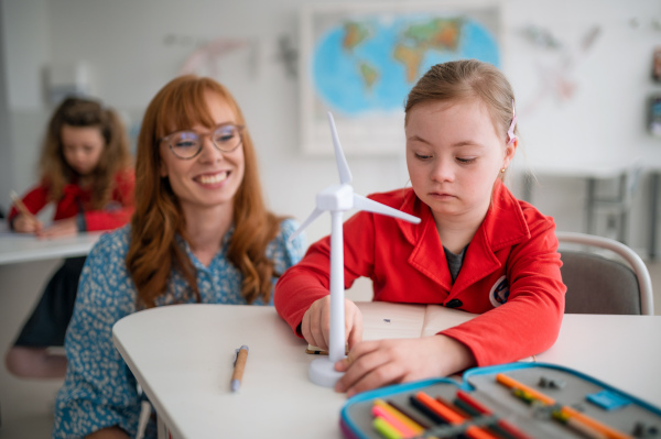 A down syndrome schoolgirl with model of wind turbine with help of teacher learning about eco-friendly renewable sources of energy in class at school, integration concept.