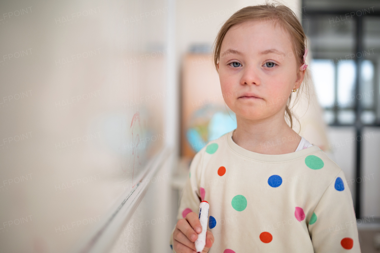 A little schoolgirl with down syndrome standing in front of whiteboard and looking at camera