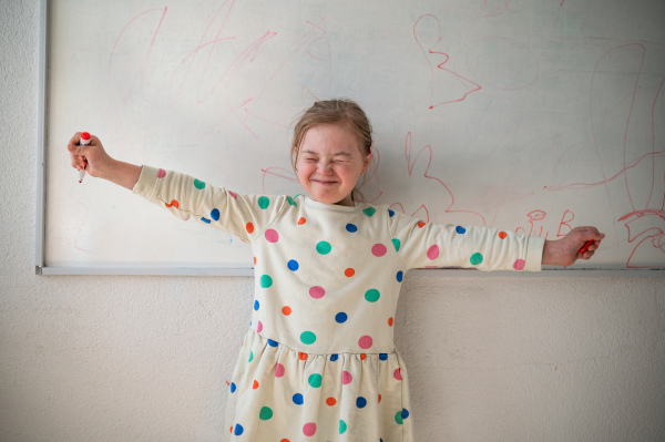 A happy little schoolgirl with down syndrome standing in front of whiteboard and looking at camera