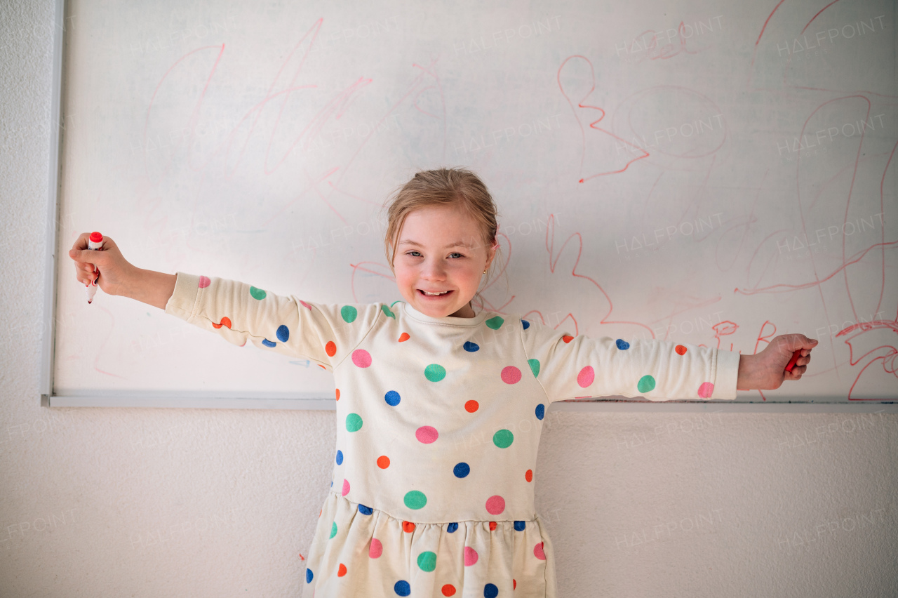 A happy little schoolgirl with down syndrome standing in front of whiteboard and looking at camera