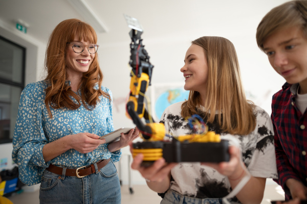 Cheerful students showing their builded robotic toy to a young science teacher at robotics classroom at school.