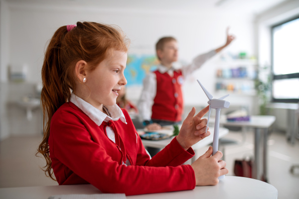 A schoolgirl holding wind turbine and learning about eco-friendly renewable sources of energy in classroom at school