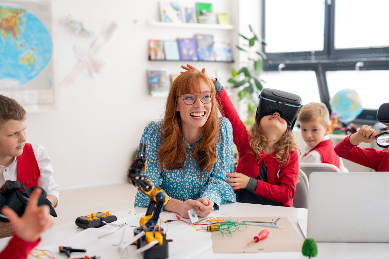 Happy schoolgirl wearing virtual reality goggles at class, having fun with teacher and her classmates.