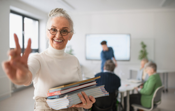 A happy senior woman student with books raising hand and looking at camera in classroom.