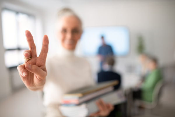 A happy senior woman student with books raising hand and looking at camera in classroom.