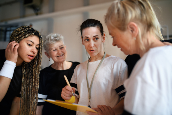 A female sport coach with clipboard discussing tactics with young and old women team training for match in gym.