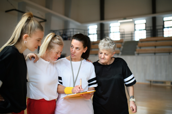 A female sport coach with clipboard discussing tactics with young and old women team training for match in gym.