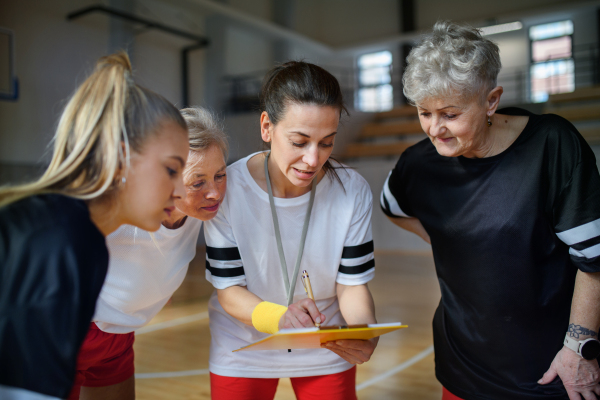 A female sport coach with clipboard discussing tactics with young and old women team training for match in gym.