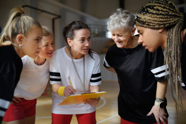 A female sport coach with clipboard discussing tactics with young and old women team training for match in gym.