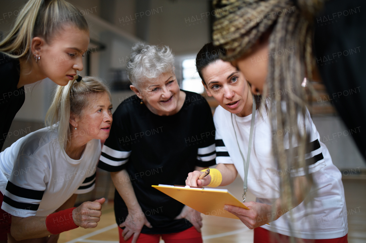 A female sport coach with clipboard discussing tactics with young and old women team training for match in gym.