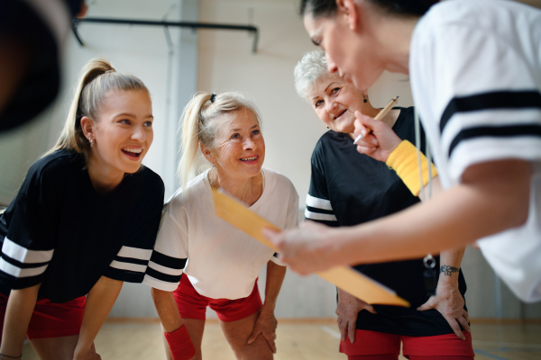 A female sport coach with clipboard discussing tactics with young and old women team training for match in gym.