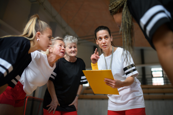 A female sport coach with clipboard discussing tactics with young and old women team training for match in gym.