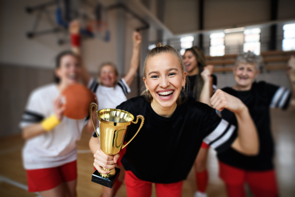 A group of young and old women, basketball team players, in gym with trophy celebrating victory.