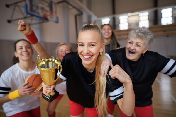 A group of young and old women, basketball team players, in gym with trophy celebrating victory.