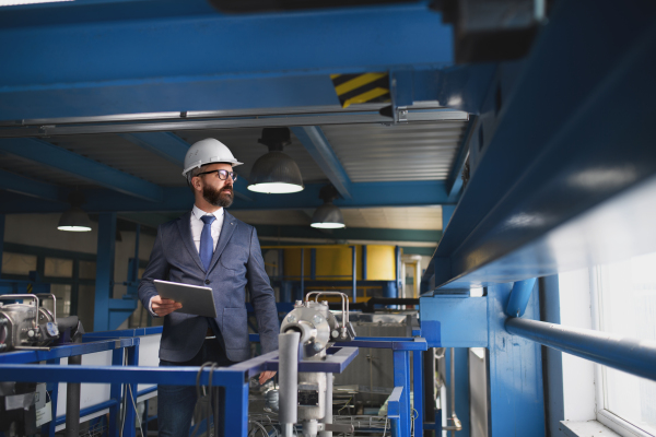 A chief Engineer in the hard hat walks through industrial factory while holding tablet.