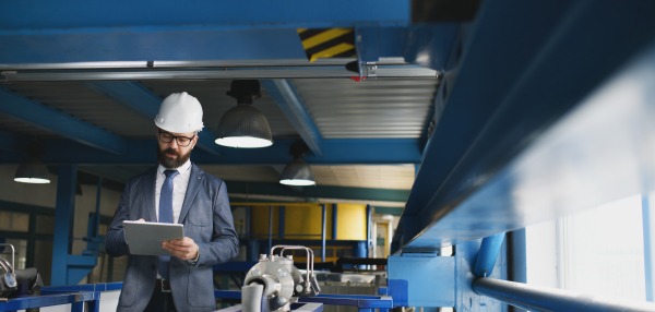 A chief Engineer in the hard hat walks through industrial factory while holding tablet.