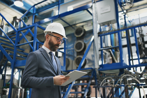 A chief Engineer in the hard hat walks through industrial factory while holding tablet.